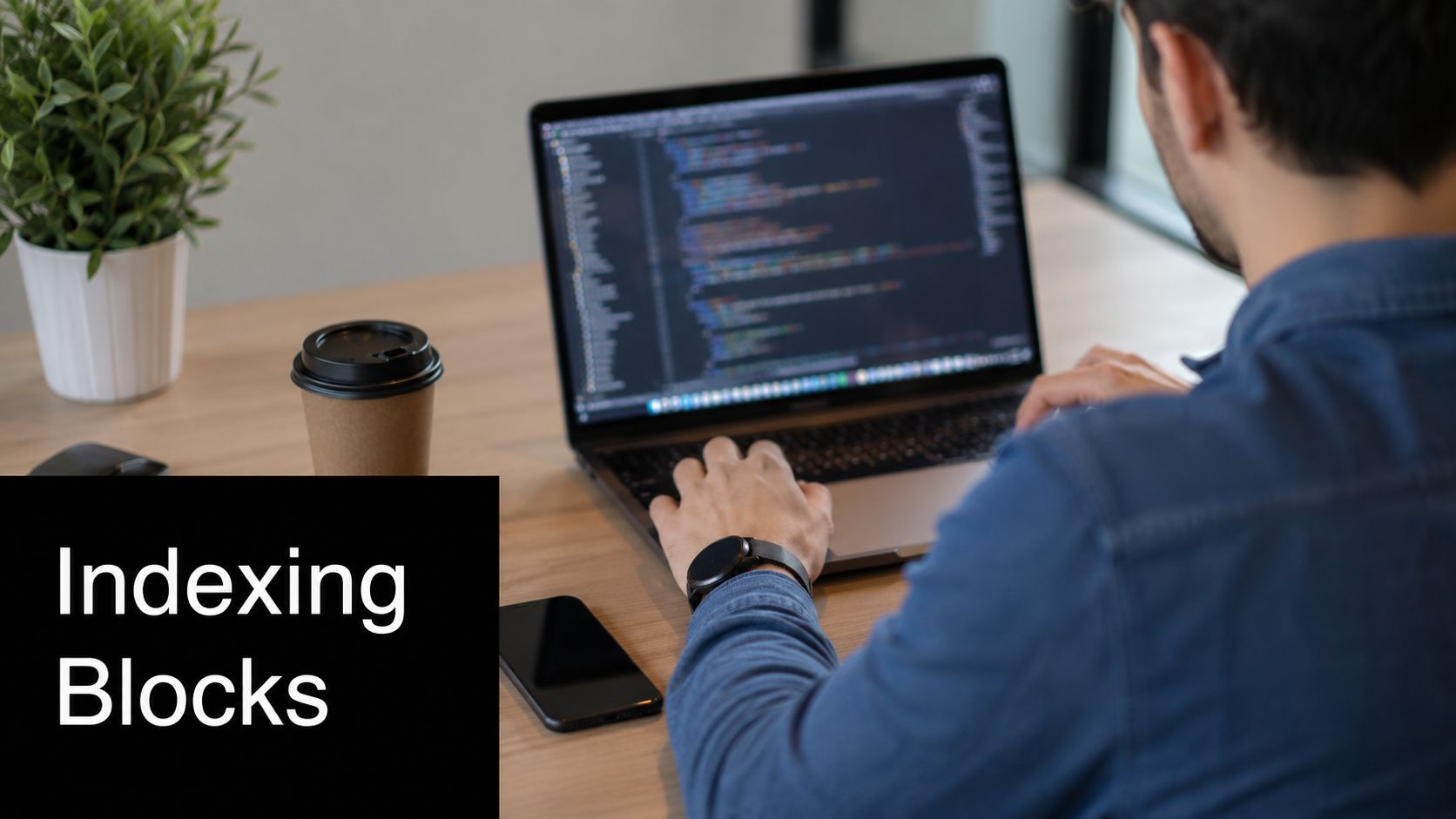 A male programmer working on a laptop at a wooden desk with a coffee cup.